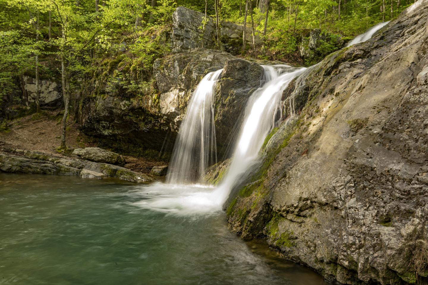 Waterfall flowing over rocks into a clear, green pool surrounded by lush greenery.