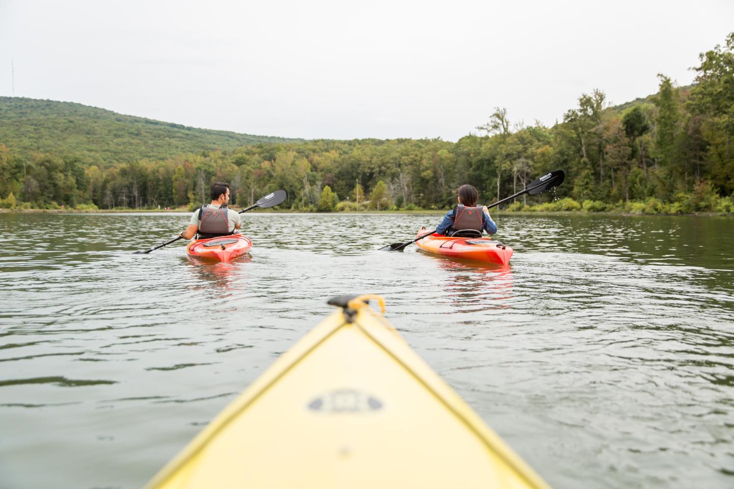 Two people kayaking on a lake surrounded by forested hills.