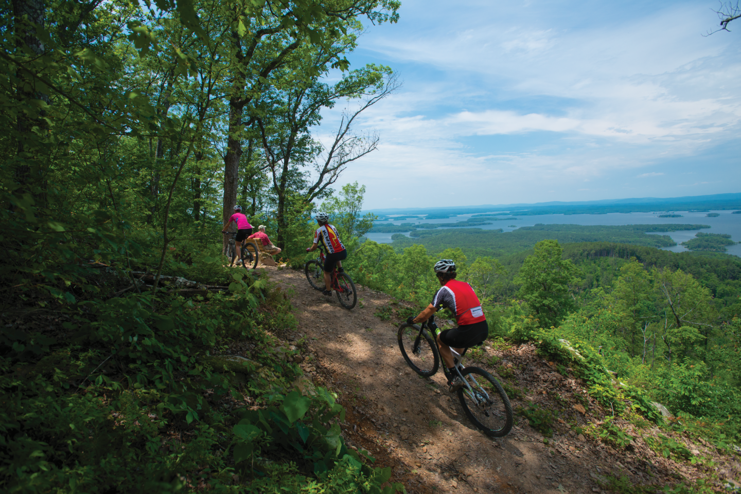 Cyclists ride a forested trail overlooking a scenic lake view.