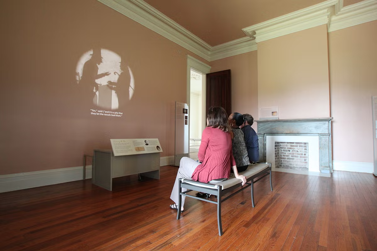 Two people sit on a bench in a small museum room with a fireplace.