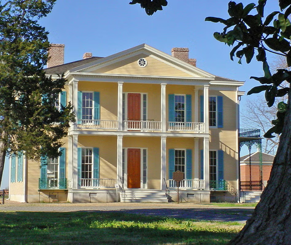 Historic yellow house with columns, green shutters, and a red door, surrounded by trees.