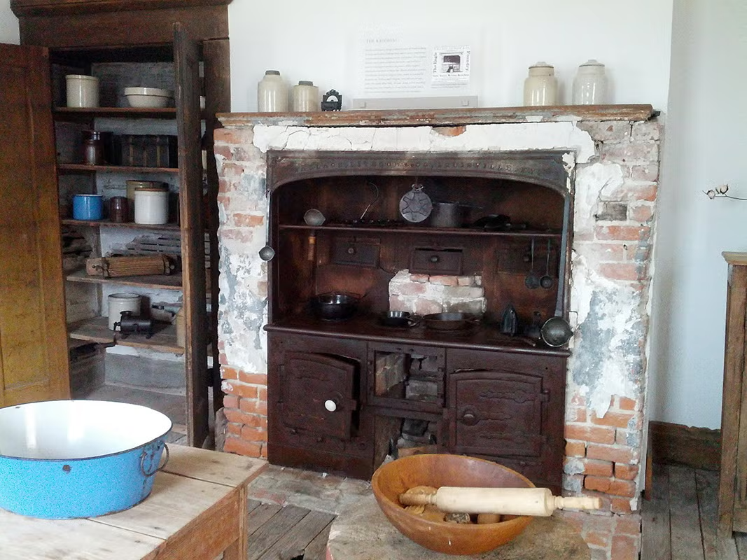 Historic kitchen with brick stove, wooden shelves, and baking items.