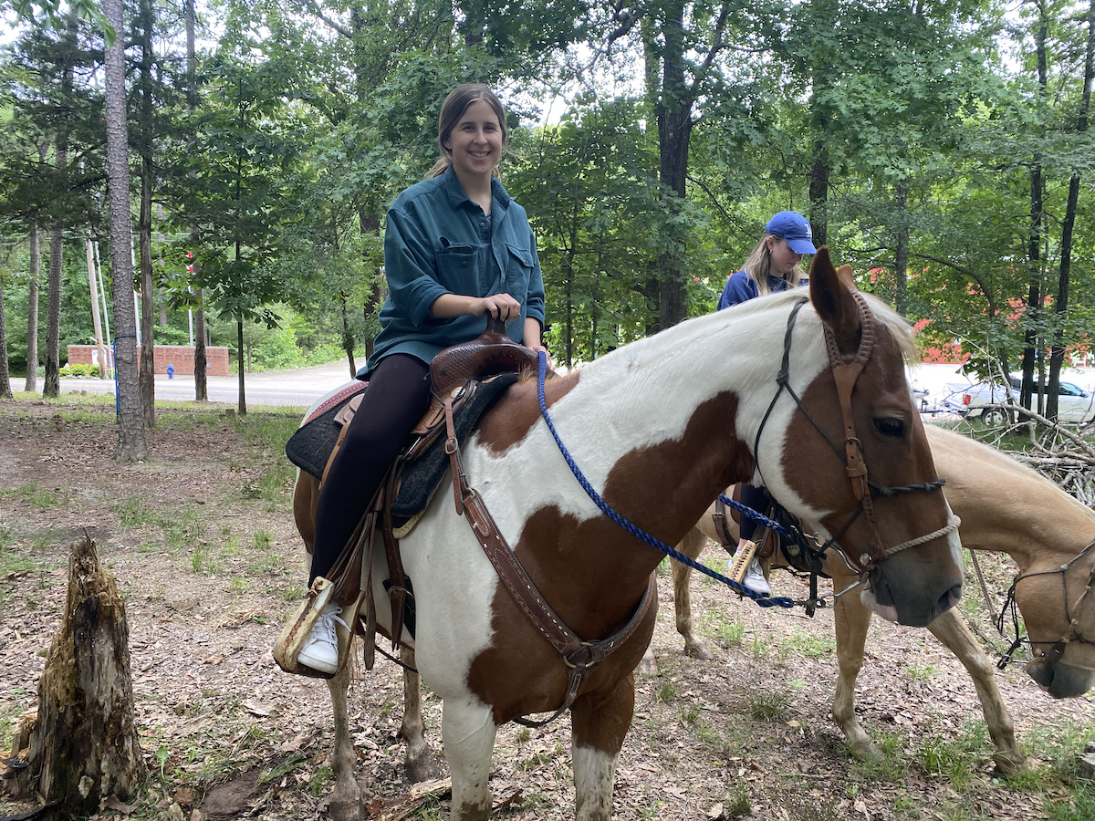 Woman smiling while riding a brown and white horse in a wooded area.