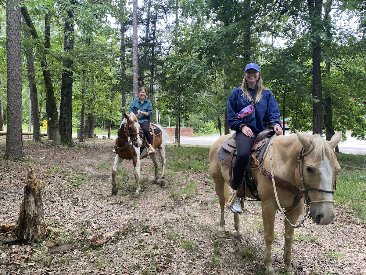 Two people riding horses in a wooded area with trees and grass.