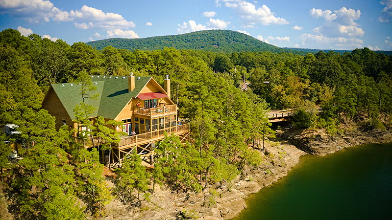 Rustic cabin by a lake, surrounded by trees with a mountain backdrop.