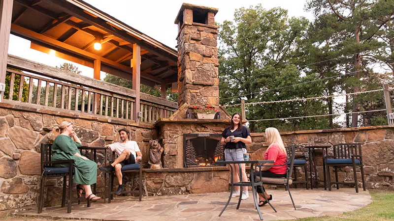 Outdoor patio with four people seated near a stone fireplace, surrounded by trees.