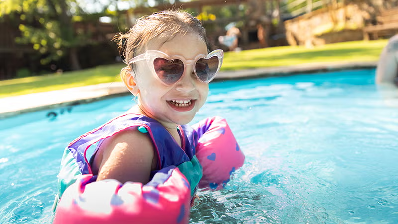 Child in a pool with heart-shaped sunglasses and colorful floaties, smiling in sunlight.