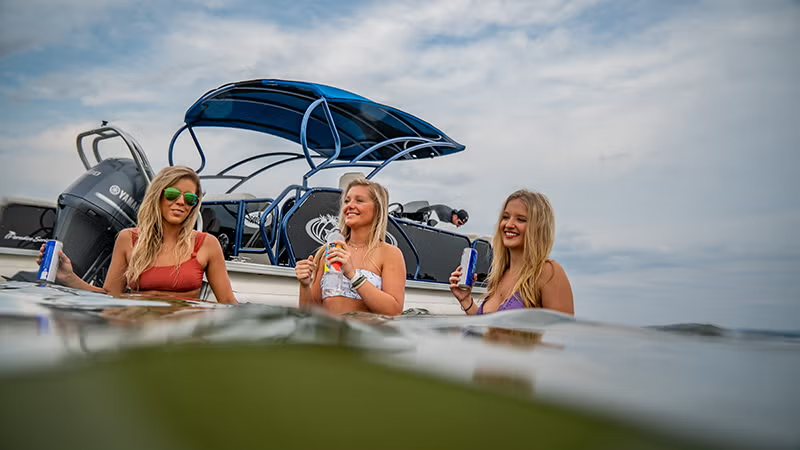 Three women in swimsuits enjoy drinks near a boat on the water.