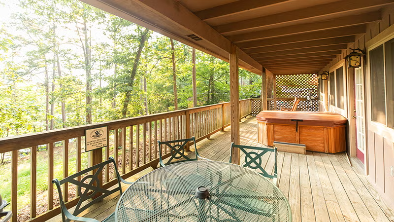 Covered wooden deck with a glass table, chairs, and a hot tub overlooking trees.