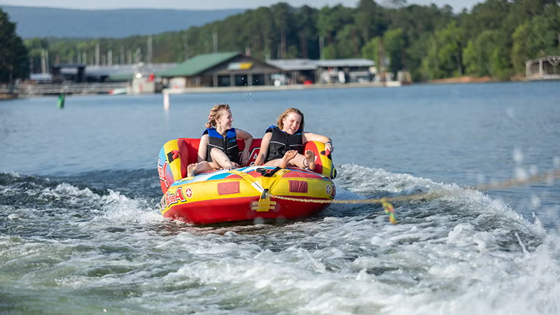 Two people tubing on a lake, smiling, with trees and a building in the background.