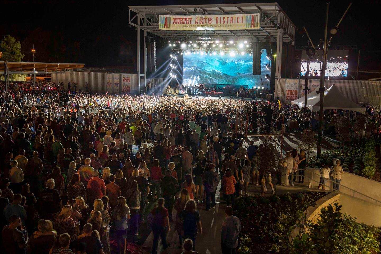 Crowd at night concert with lit stage and large screen.