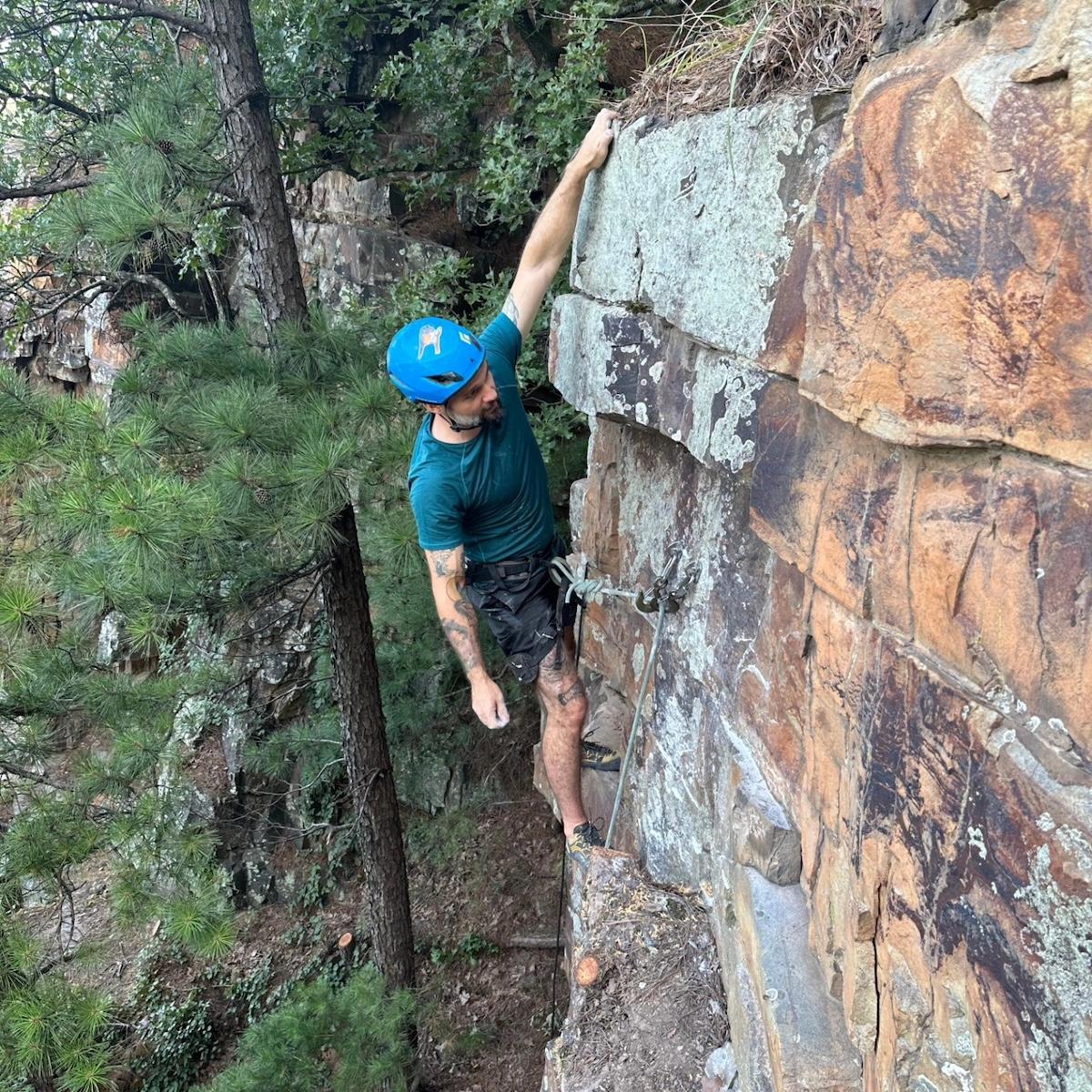 Rock climber in blue helmet scaling a cliff with trees in the background.
