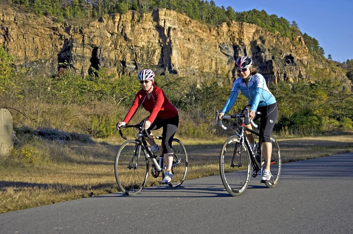 Two cyclists ride on a road against a rocky cliff backdrop.