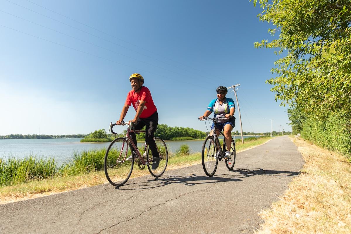 Two people cycling on a lakeside path under a clear blue sky.
