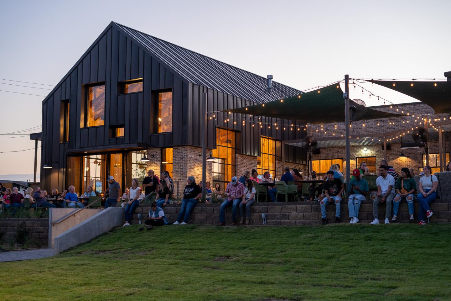 Modern barn-style building with people gathered on a grassy lawn at dusk.