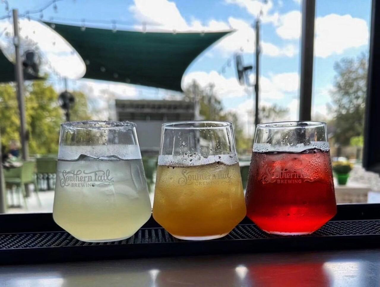 Three cocktails on a bar, each a different color, with an outdoor patio view.