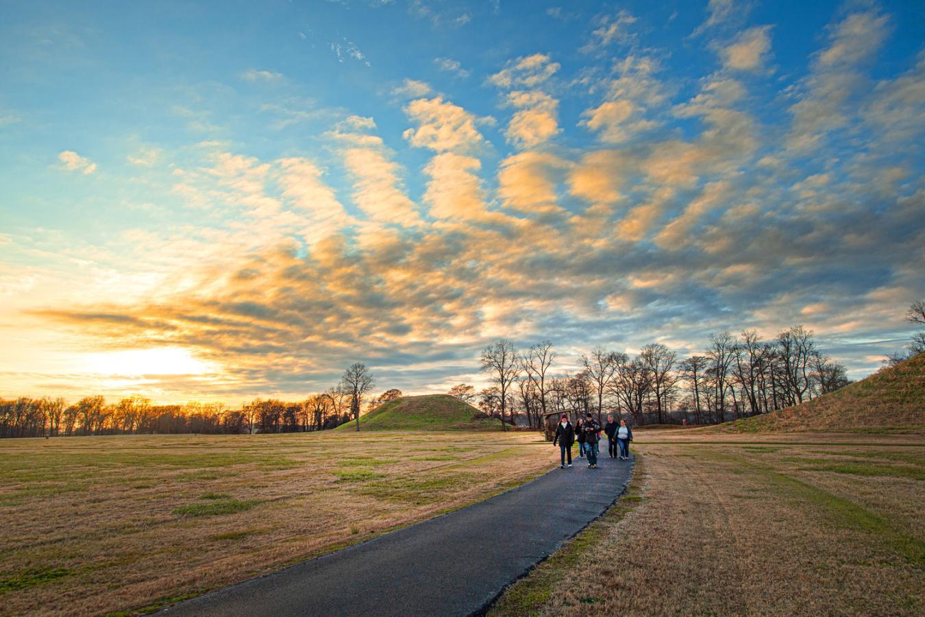 Path through field, people walking, sunset with scattered clouds.