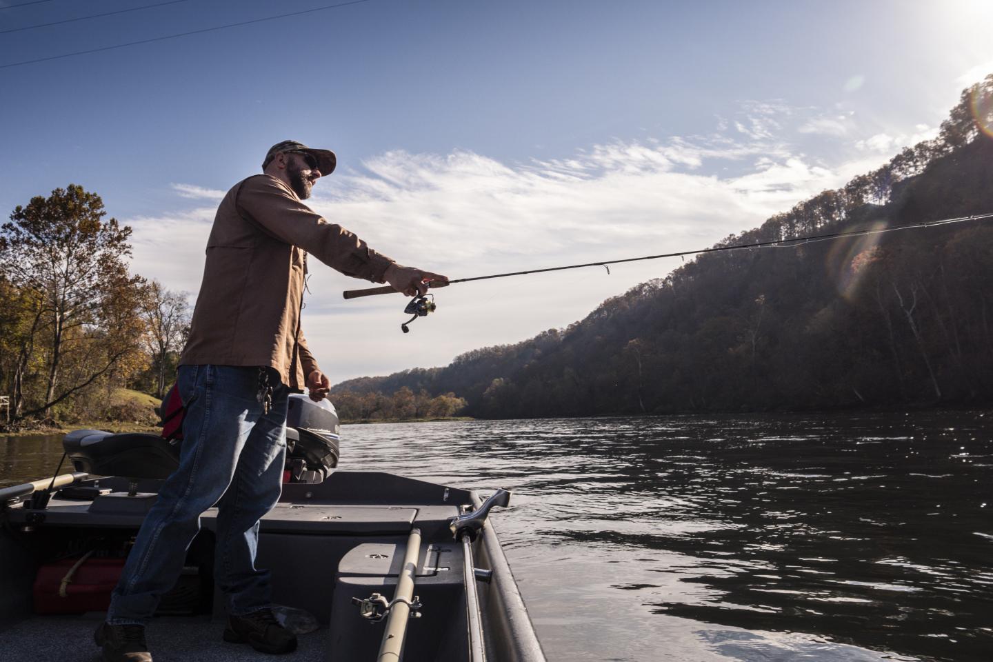 Man fishing on a boat in a river, surrounded by hills and trees.