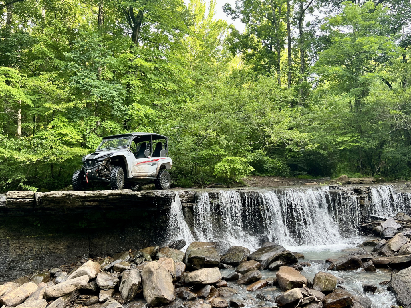 Off-road vehicle on a stone bridge over a small waterfall in lush forest.