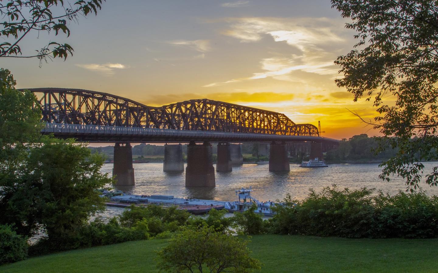 Bridge over a river at sunset with trees and grass in the foreground.