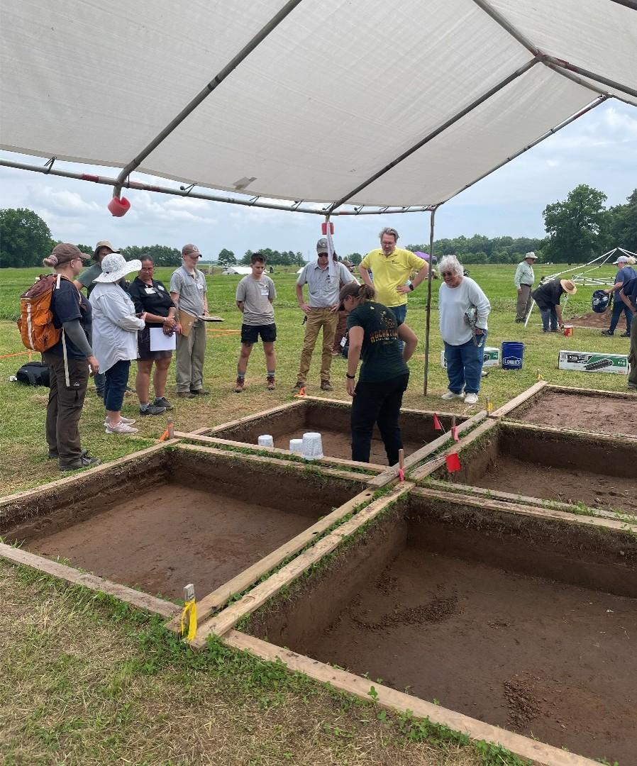 Archaeologists examine dig sites under a large canopy in a grassy field.