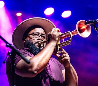 Trumpet player in a hat performs on stage with colorful lighting.
