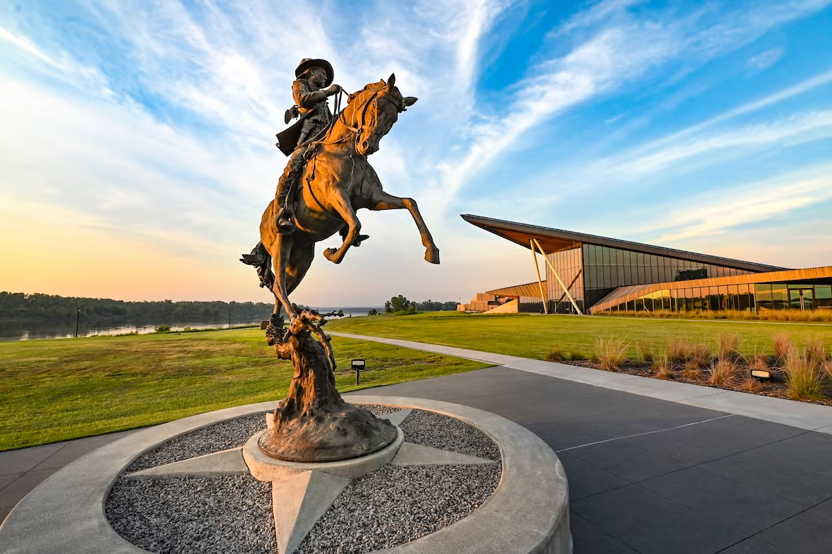 Bronze horse and rider statue under a blue sky with clouds, near a modern building.