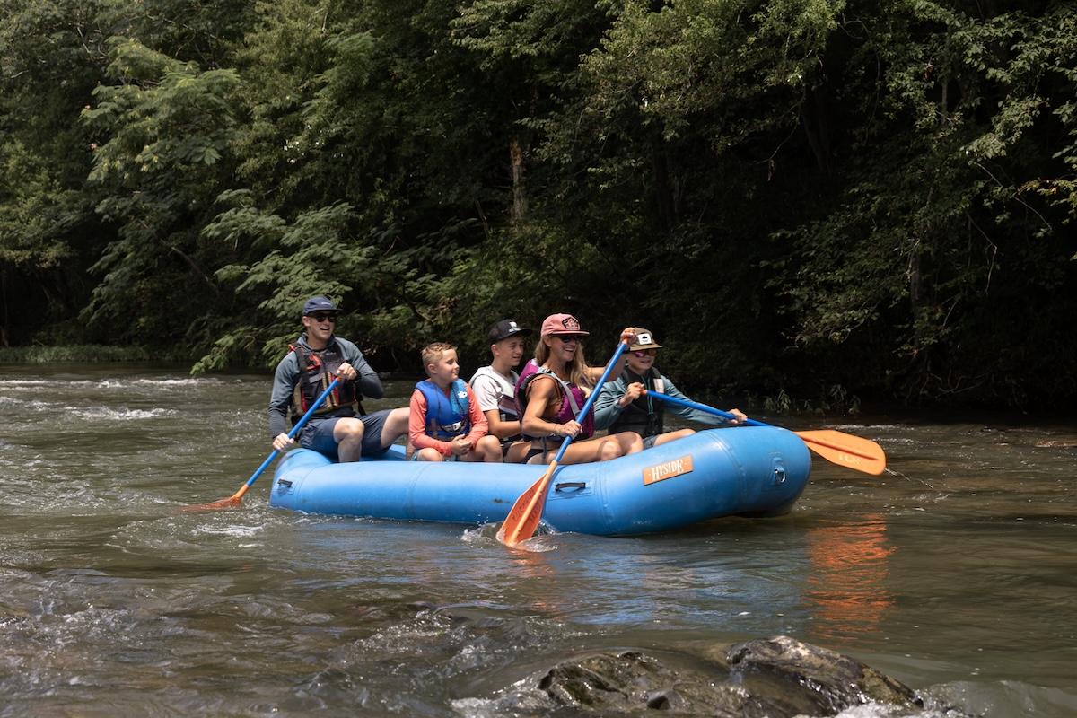 Group of people rafting on a river in a blue inflatable boat, surrounded by trees.