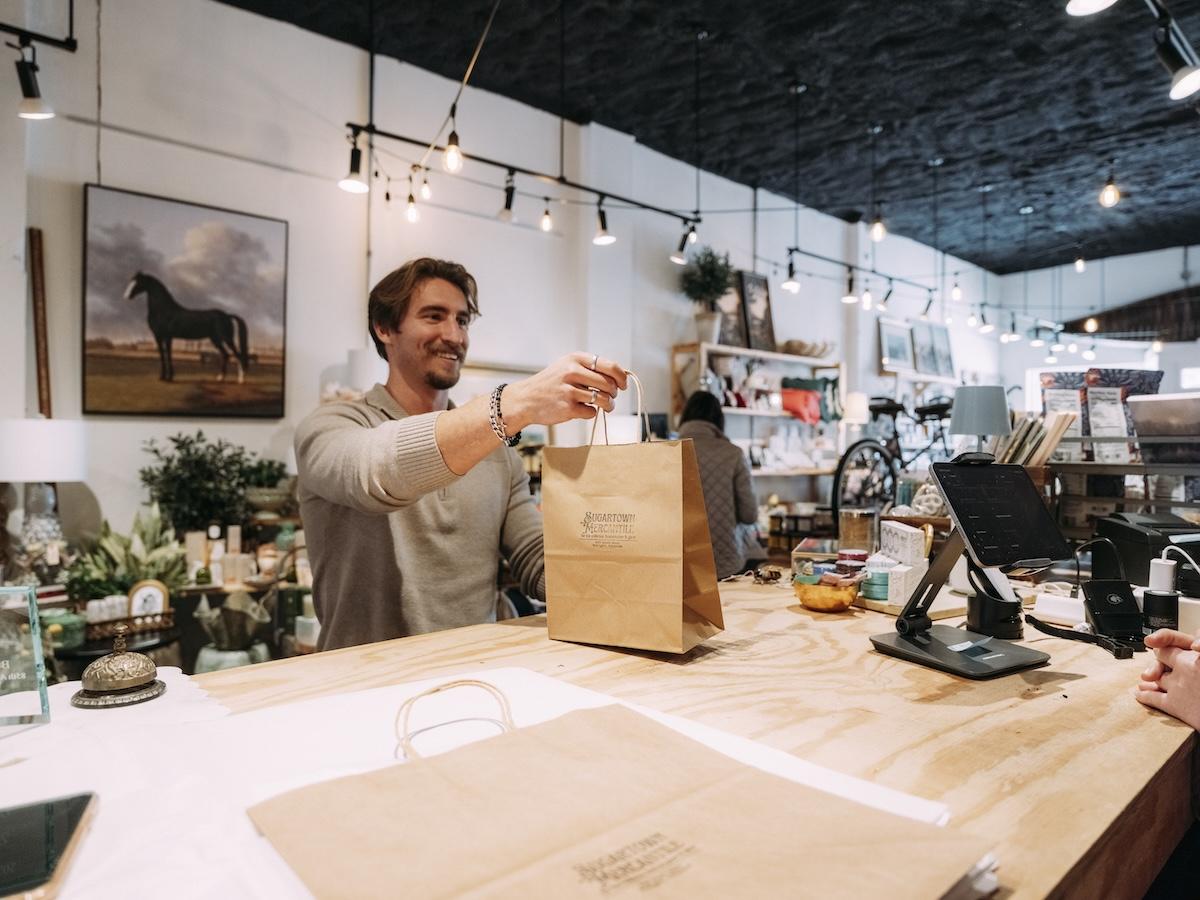 Man handing a paper bag to a customer at a shop counter.