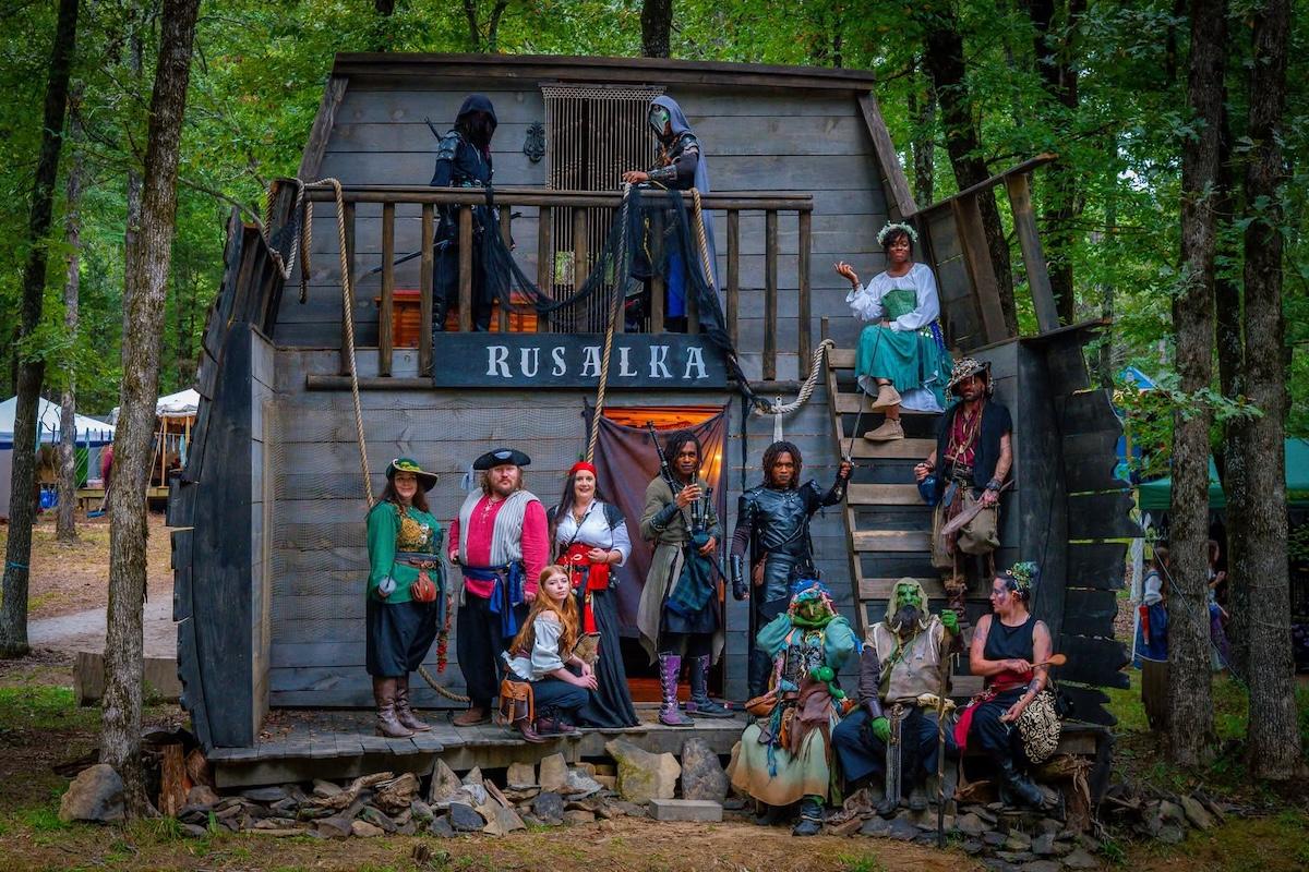 Medieval-dressed group outside a rustic wooden building in a forest setting.