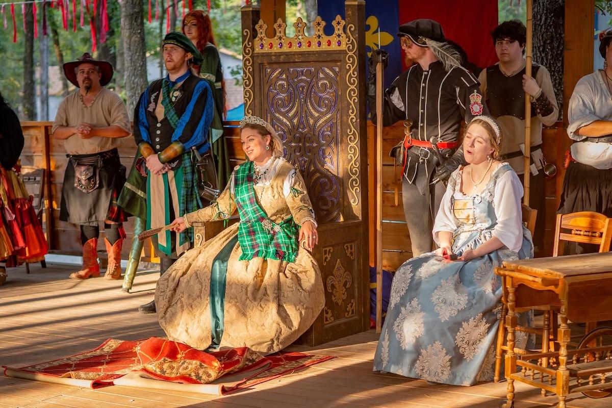 Renaissance fair scene with people in period costumes, woman on a throne, others standing nearby.