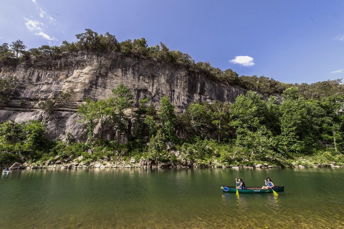 Canoe on a river with rocky cliff and trees under blue sky.