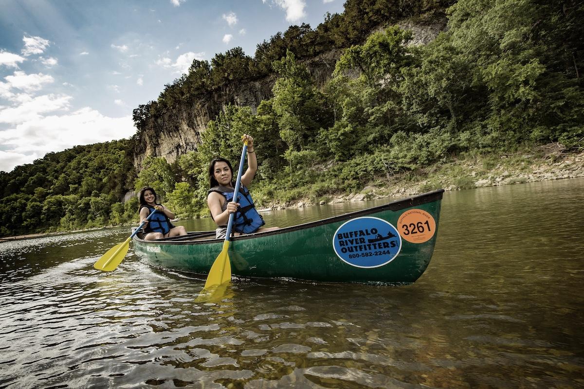 Two people canoeing on a river, surrounded by trees and cliffs.