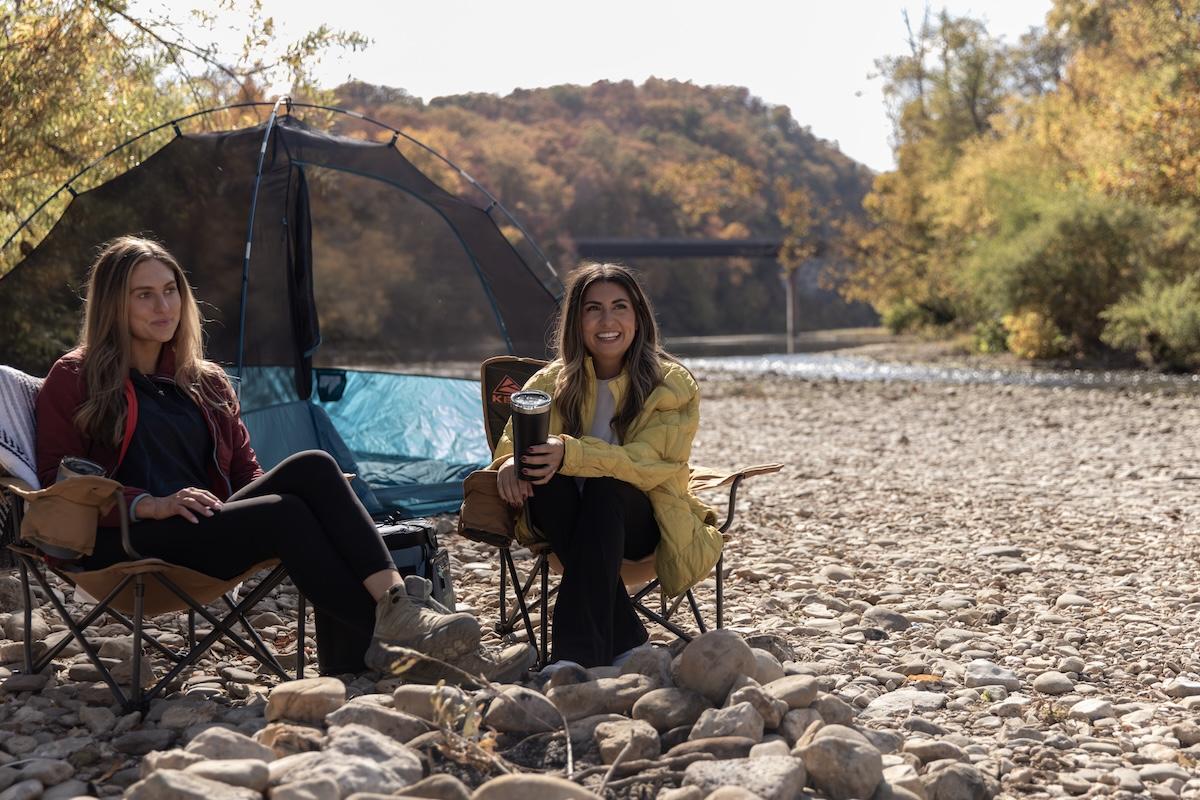 Two people relaxing in camping chairs by a rocky riverbank in autumn.