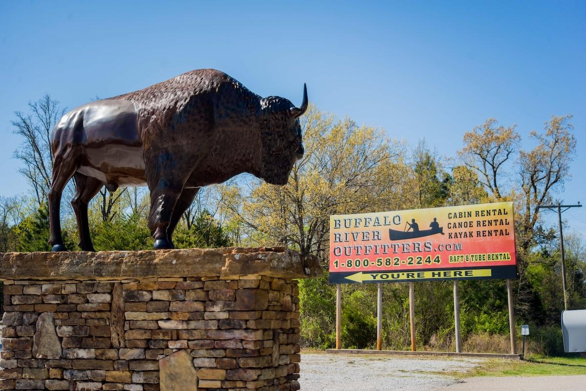 Bison sculpture on stone pedestal near a sign, clear blue sky.