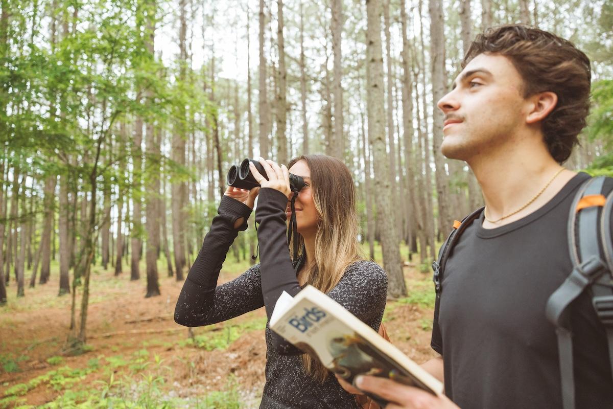 Two people hiking in a forest, one with binoculars, the other holding a book.