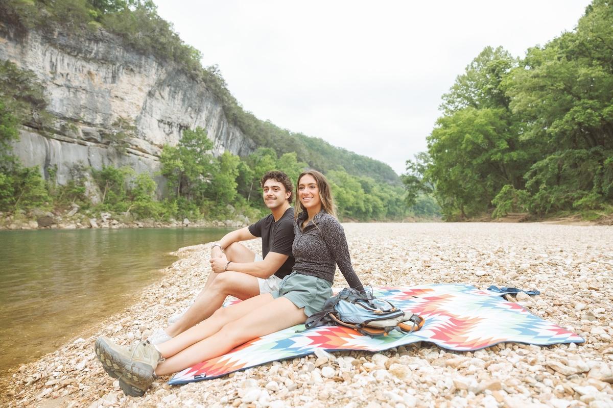Couple sitting on a colorful blanket by a river, surrounded by trees and cliffs.