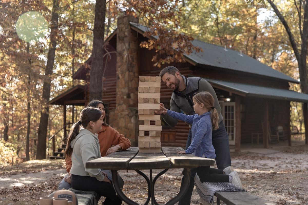 Family playing Jenga at a picnic table in autumn near a log cabin.