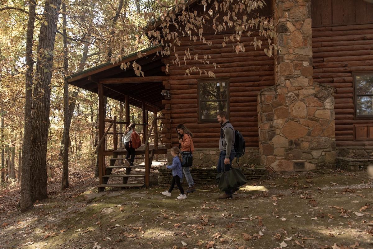 Family walking past a rustic log cabin in a wooded area.