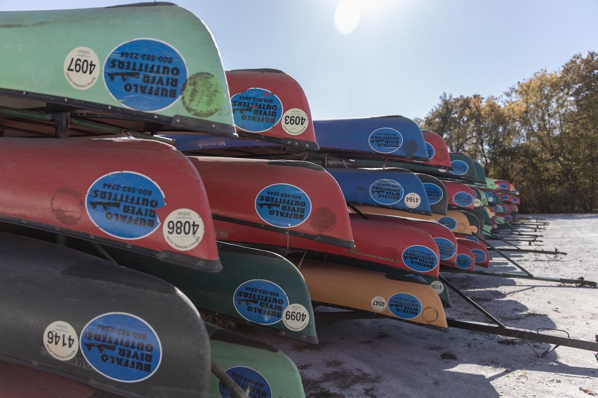 Stacked colorful canoes on a sunny day, trees in the background.