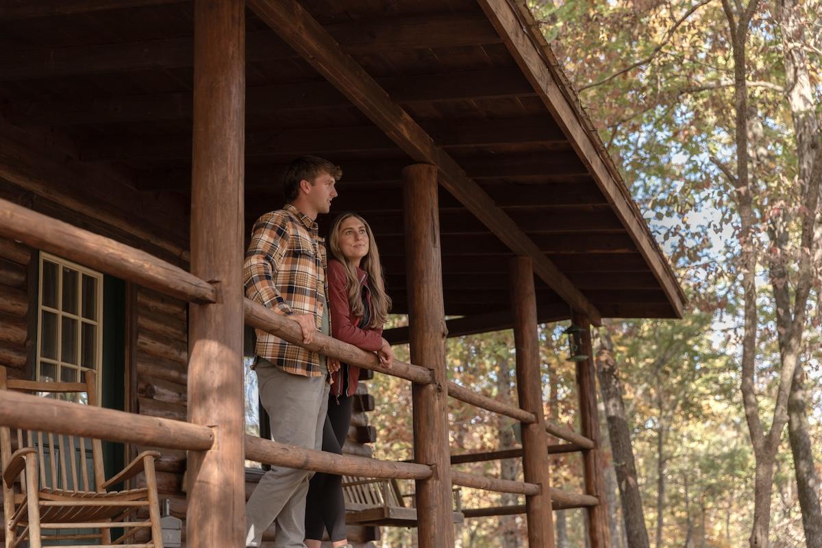 Couple standing on a rustic cabin porch in autumn woods.