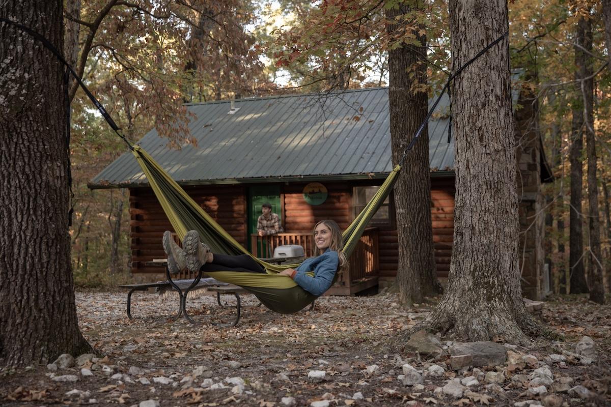 Person relaxing in a hammock near a log cabin surrounded by autumn trees.