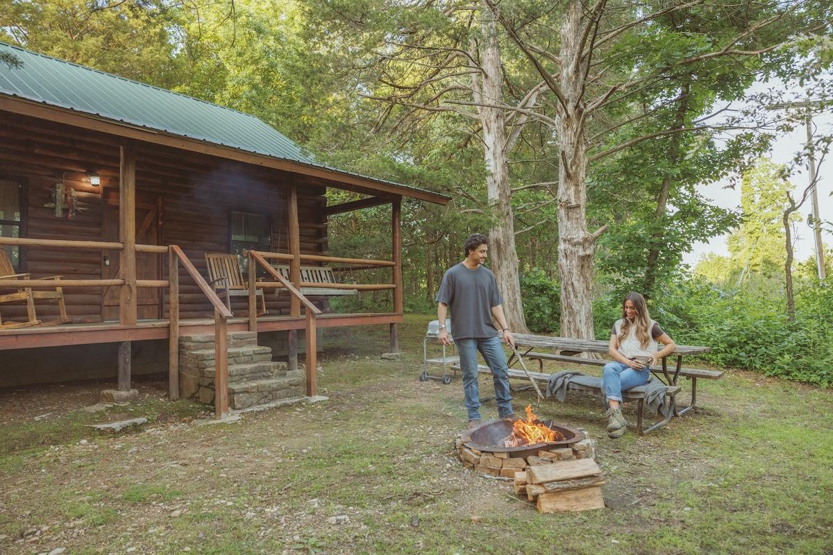 Cabin in the woods with two people by a campfire on a sunny day.