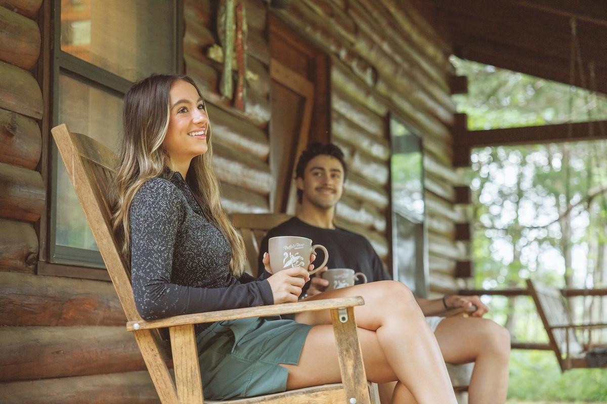 Two people smiling, sitting on a wooden porch with mugs.