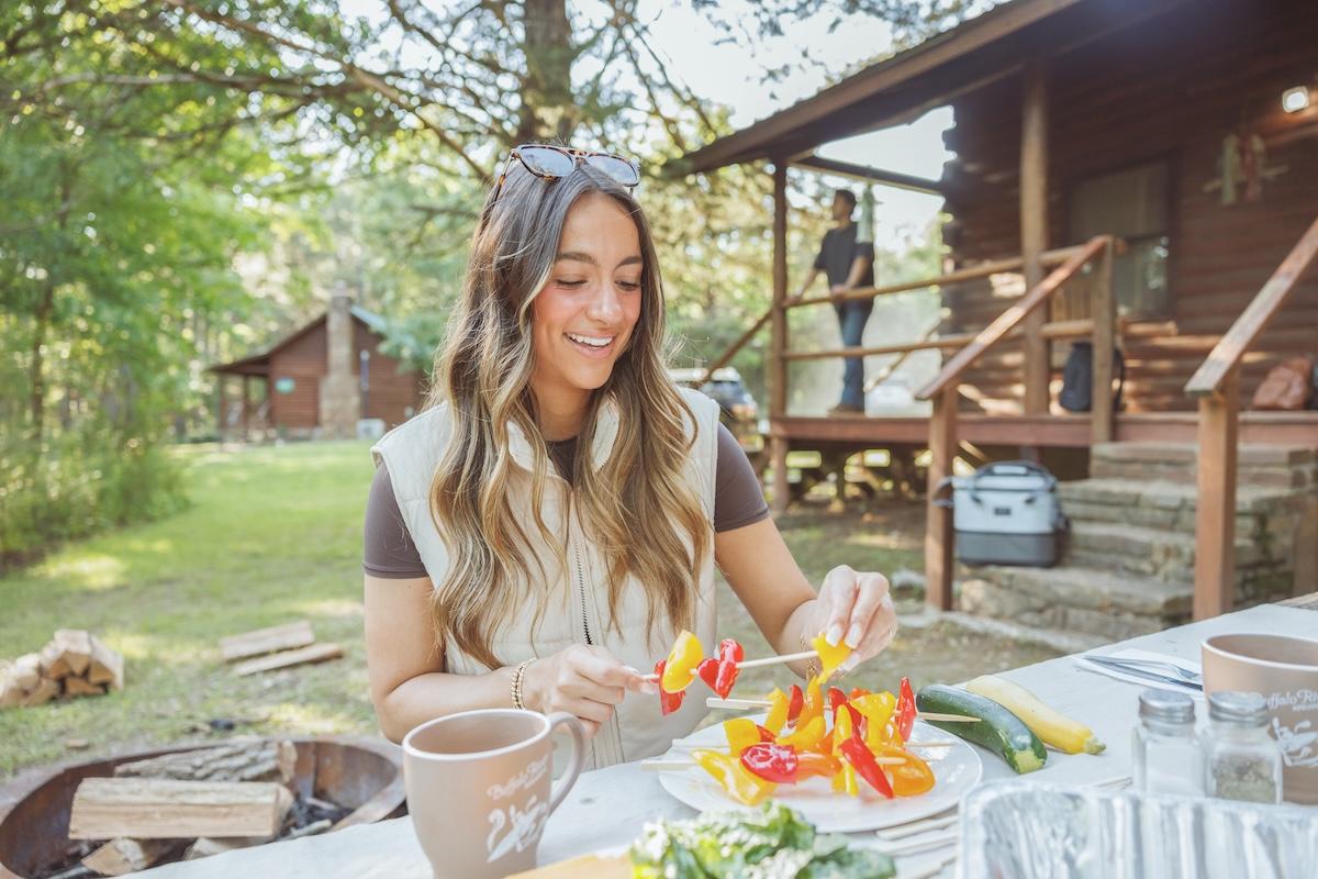 Woman preparing vegetables outdoors near a cabin in sunny weather.
