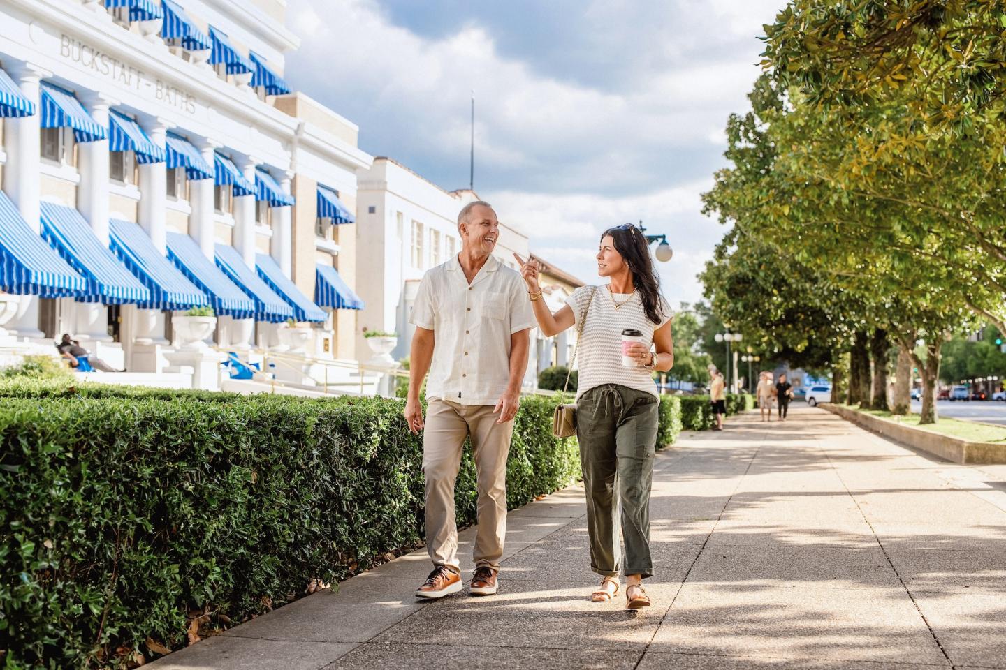 A couple walks on a sunny sidewalk beside a building with blue awnings.