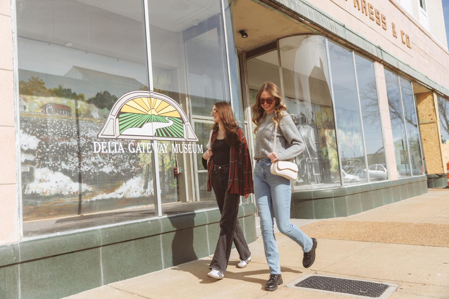 Two women walking past the Delta Gateway Museum with large windows and a colorful mural.