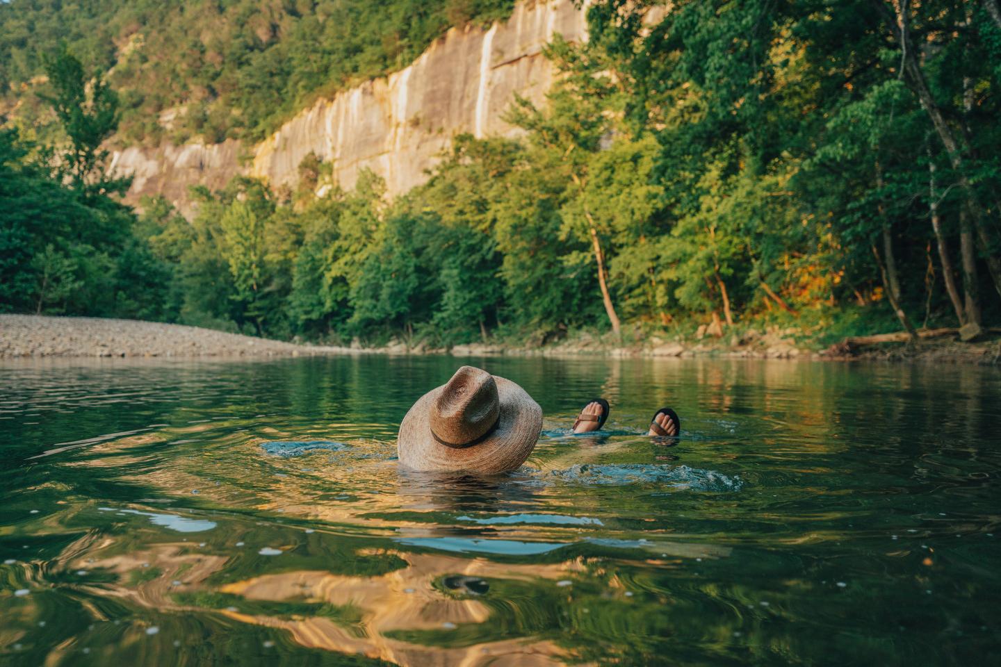 Person wearing a hat floats in a calm river surrounded by trees and cliffs.