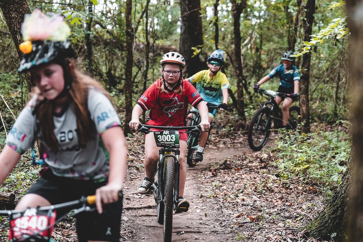 Cyclists on a forest trail, riding through leafy trees.