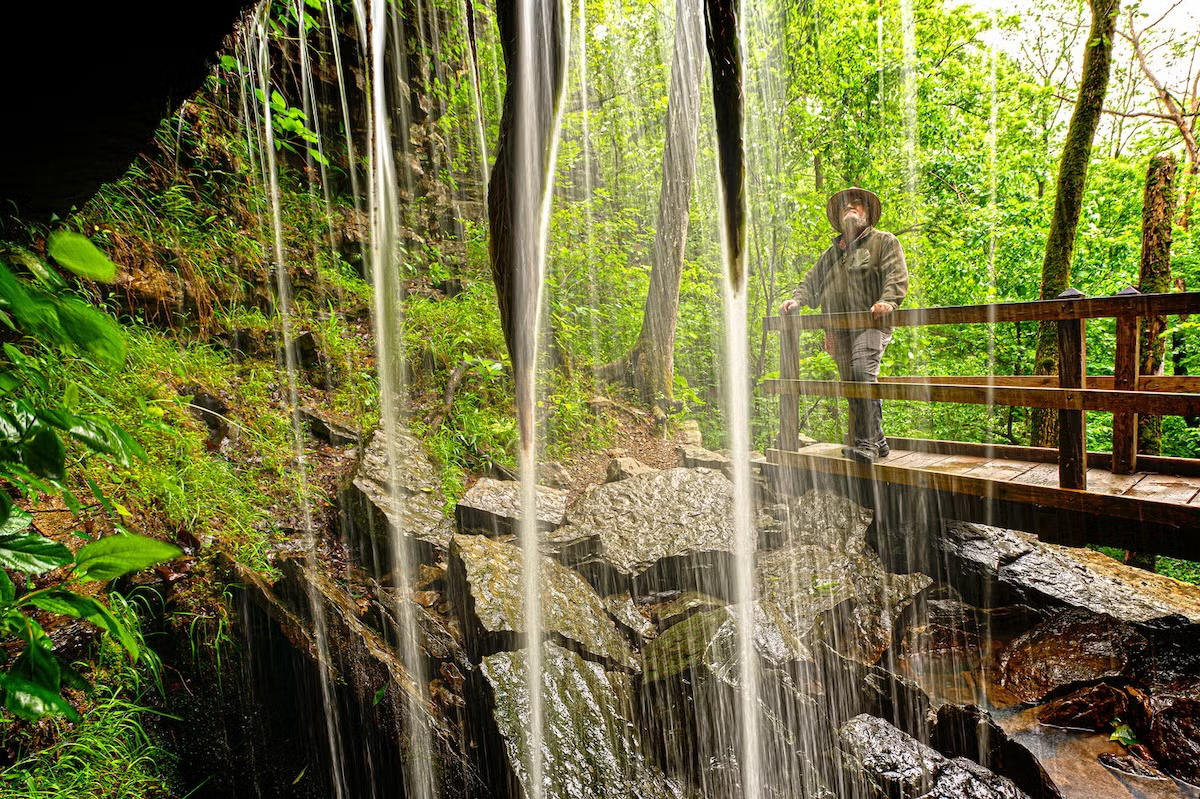 Waterfall with lush greenery and a person on a wooden bridge.